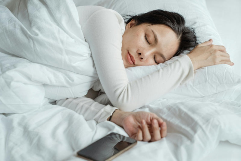 Mulher dormindo tranquilamente na cama com celular ao lado, simbolizando sono saudável.