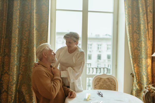 Casal sênior sorrindo em casa, sentados à mesa com comprimidos e óculos, em ambiente iluminado.