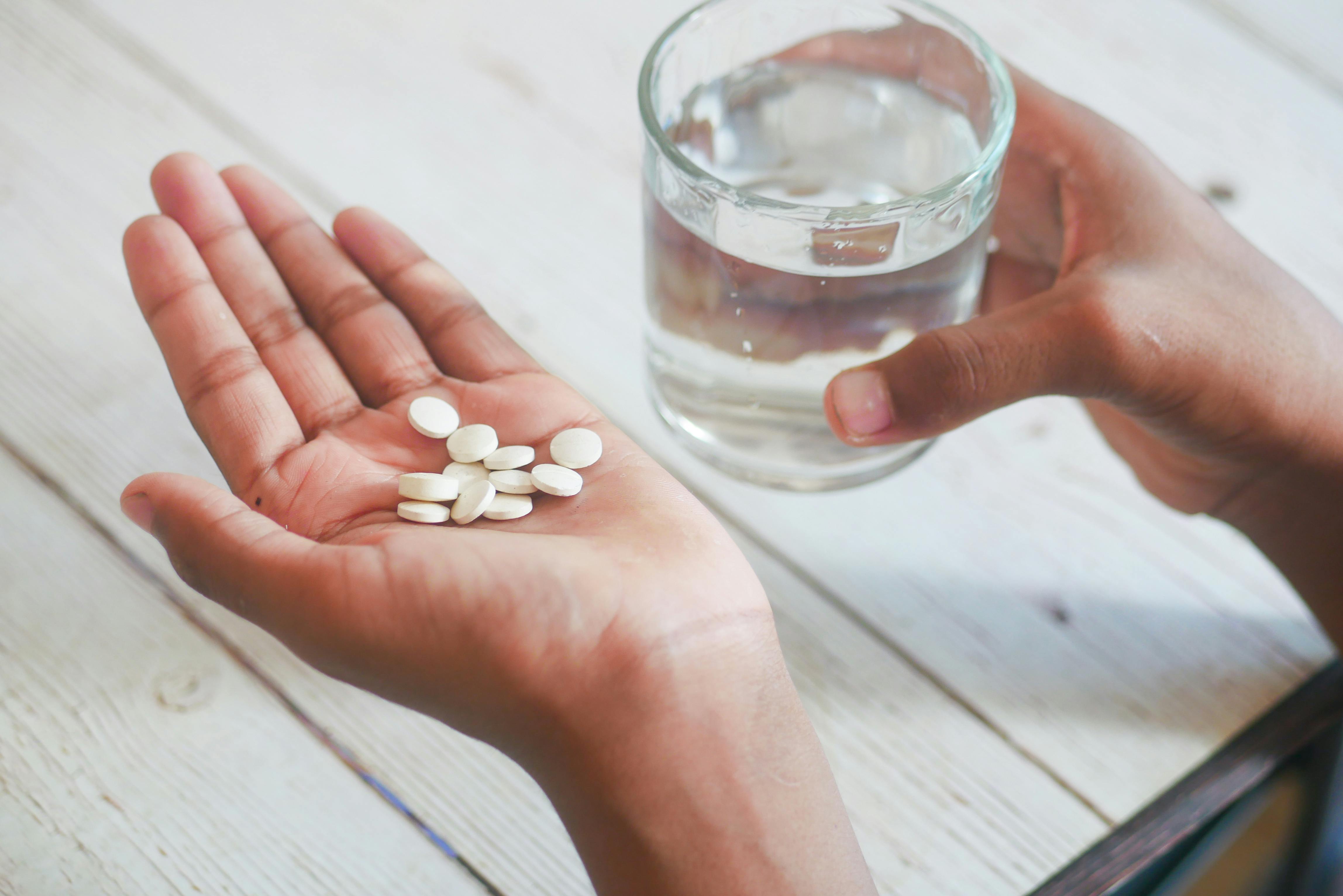 Mão com comprimidos brancos e copo de água em fundo de madeira, suplementos importados