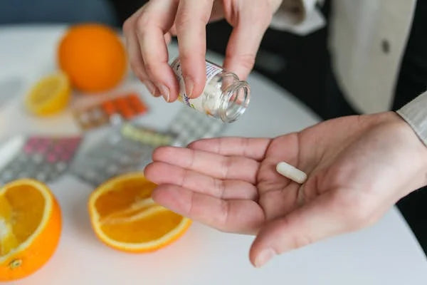 Pessoa segurando cápsula de suplemento alimentar com laranja e comprimidos ao fundo