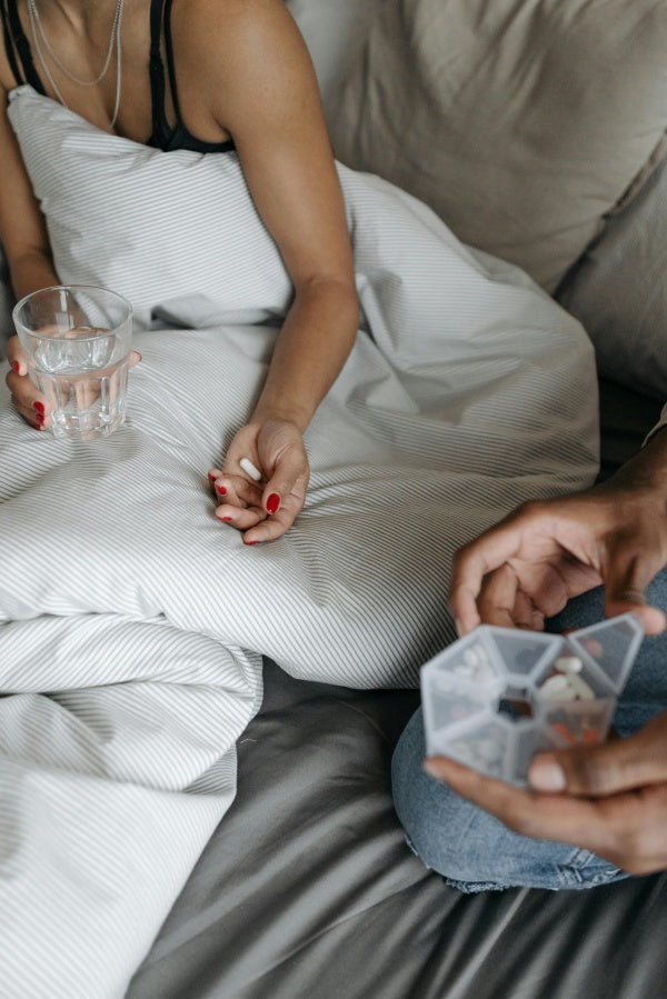 Pessoa na cama segurando copo de água e comprimido, remédios e suplementos ao lado.