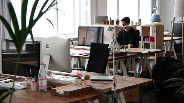 Ambiente de escritório moderno com computadores, plantas e pessoa trabalhando ao fundo.