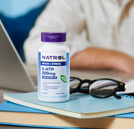 Frasco de suplemento 5-HTP Natrol 200mg sobre mesa com óculos, livros e notebook ao fundo.