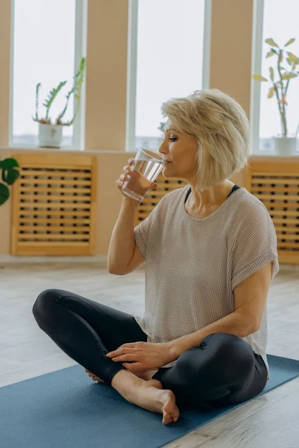 Mulher sentada no tapete de yoga, hidratando-se com copo de água, ambiente saudável