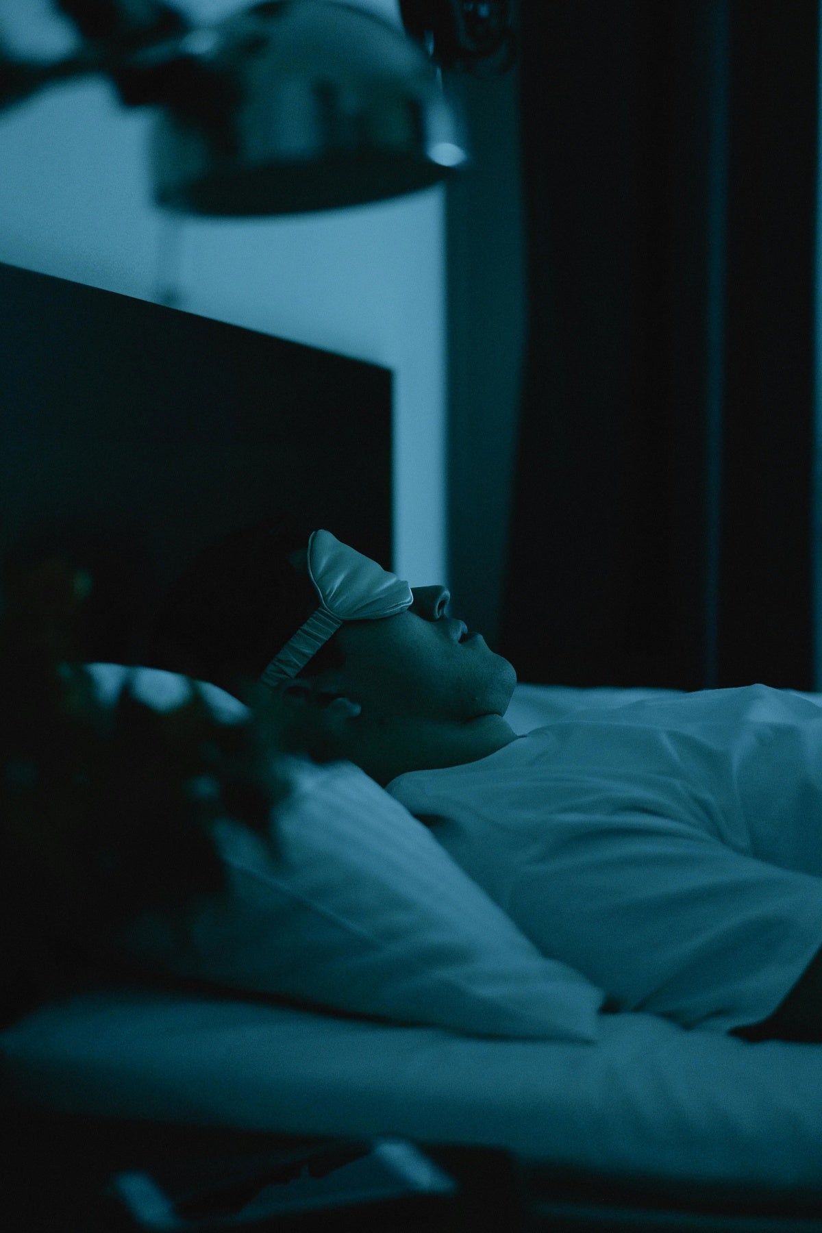 Homem dormindo em cama com máscara de dormir, quarto escuro, ambiente noturno tranquilo.