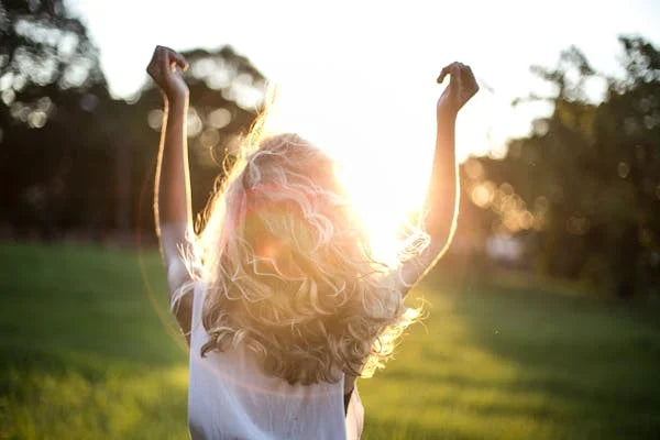 Mulher loira de braços erguidos ao ar livre ao pôr do sol, simbolizando bem-estar e vitalidade