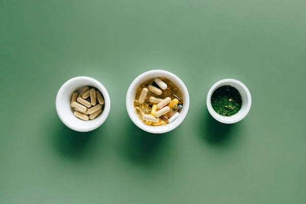 Suplementos naturais em cápsulas e ervas em potes brancos sobre fundo verde.